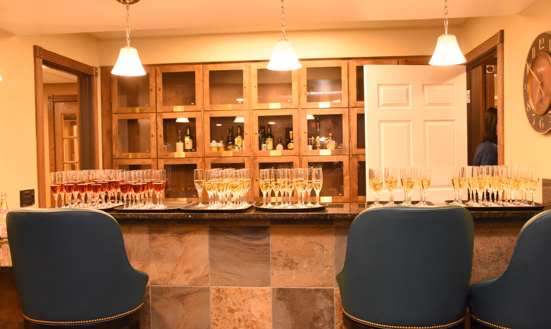 Interior view of a bar area with a countertop displaying multiple trays of filled champagne glasses. Behind the counter are wooden cabinets with glass doors containing bottles of liquor. Three pendant lights hang from the ceiling above the counter, and two blue bar stools are positioned in front. A clock is mounted on the wall to the right, and a partially open door reveals a person standing in the adjacent room.