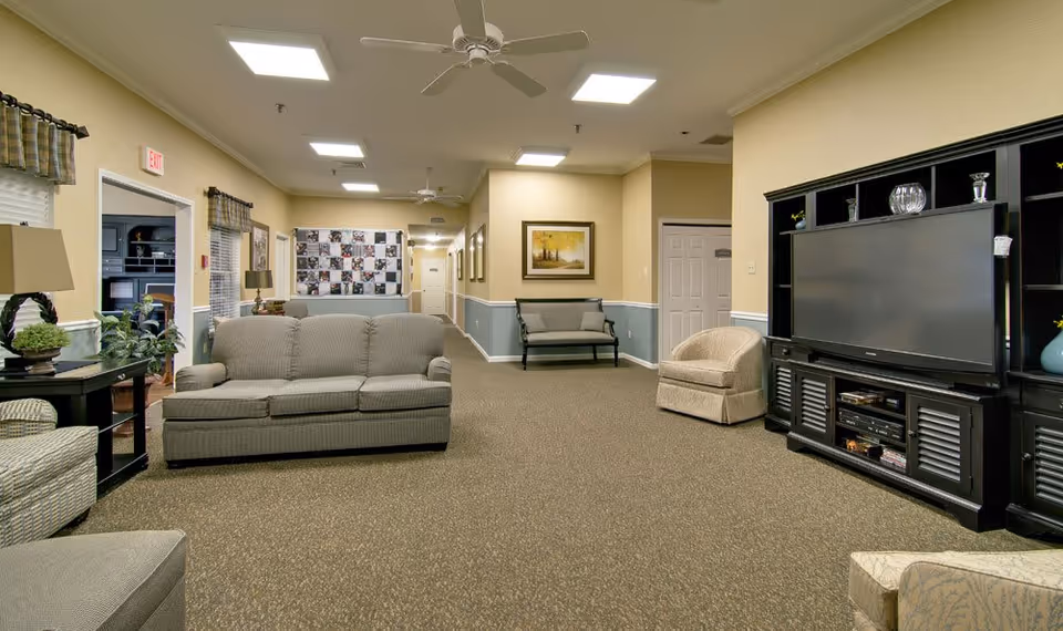 A spacious living room area in a senior living facility with beige walls and carpeted floor. The room features a gray sofa, a beige armchair, a black entertainment center with a large flat-screen TV, and a bench against the far wall. Ceiling fans and recessed lighting illuminate the space. There are framed pictures on the walls and a side table with a lamp and plants near the sofa.