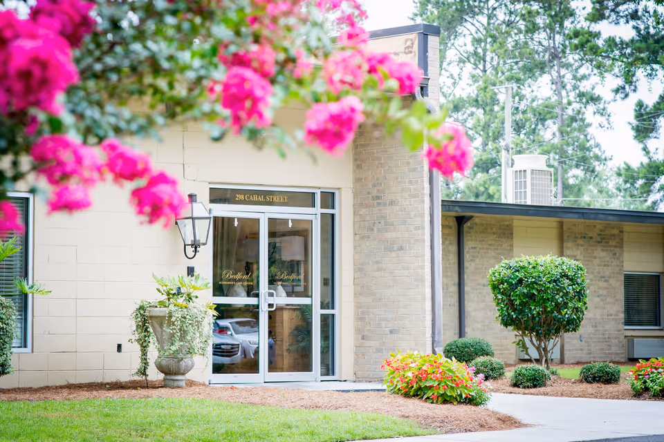 Entrance of Bedford Alzheimer's Care Center with glass double doors, beige brick walls, and a landscaped garden featuring green bushes and vibrant pink flowers in the foreground.