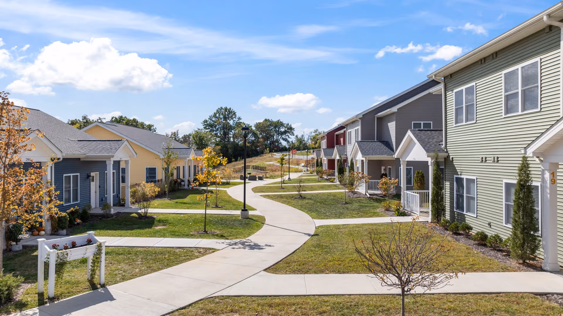 A sunny outdoor pathway winding through a senior living community with small, colorful houses on either side. The houses have porches and are surrounded by young trees and well-maintained lawns under a blue sky with scattered clouds.