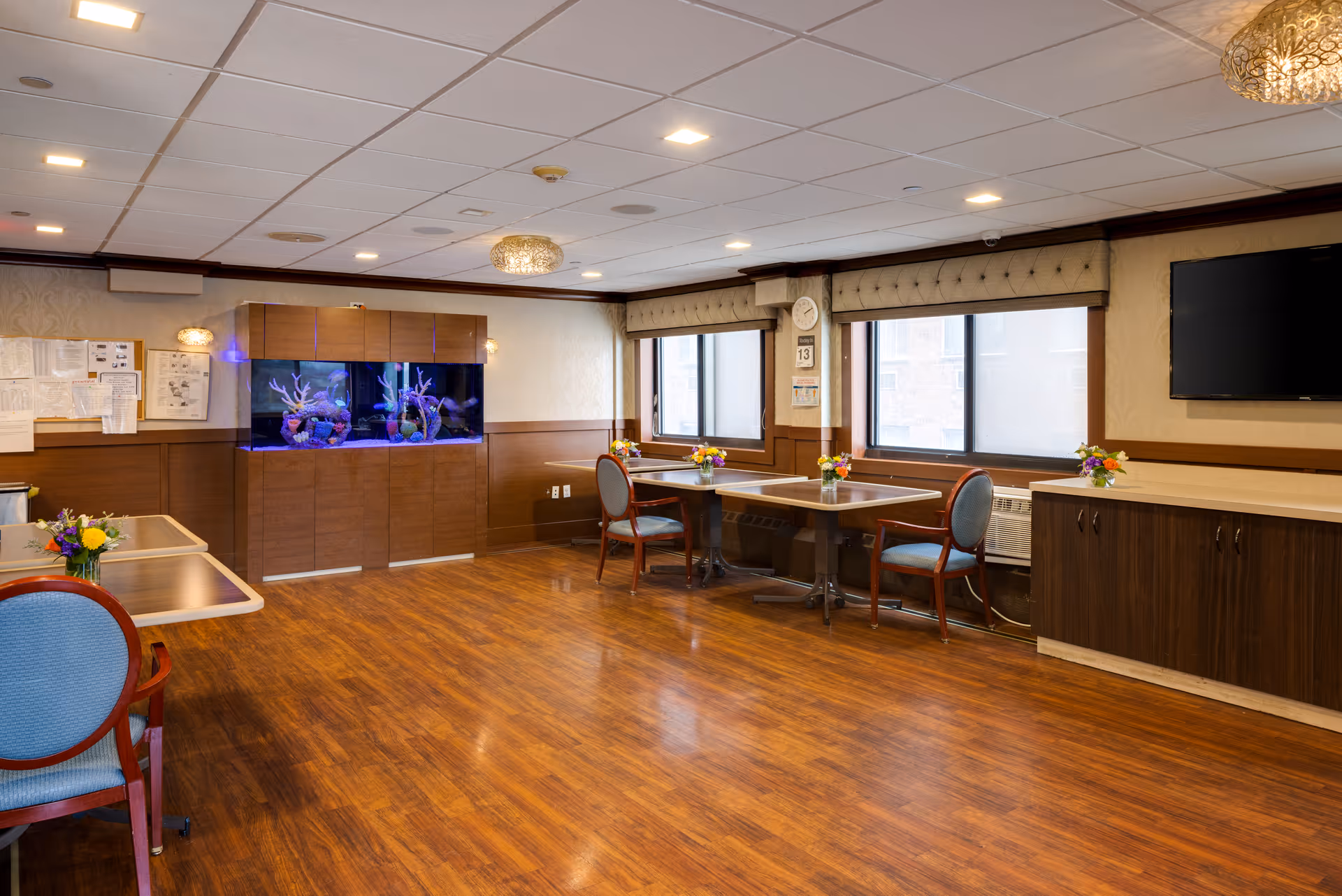 A spacious, well-lit common area in a nursing home featuring wooden flooring, several tables with chairs, small flower arrangements on the tables, a large built-in aquarium with colorful coral decorations, two large windows with beige valances, a wall-mounted flat screen TV, and a clock and calendar on the wall.