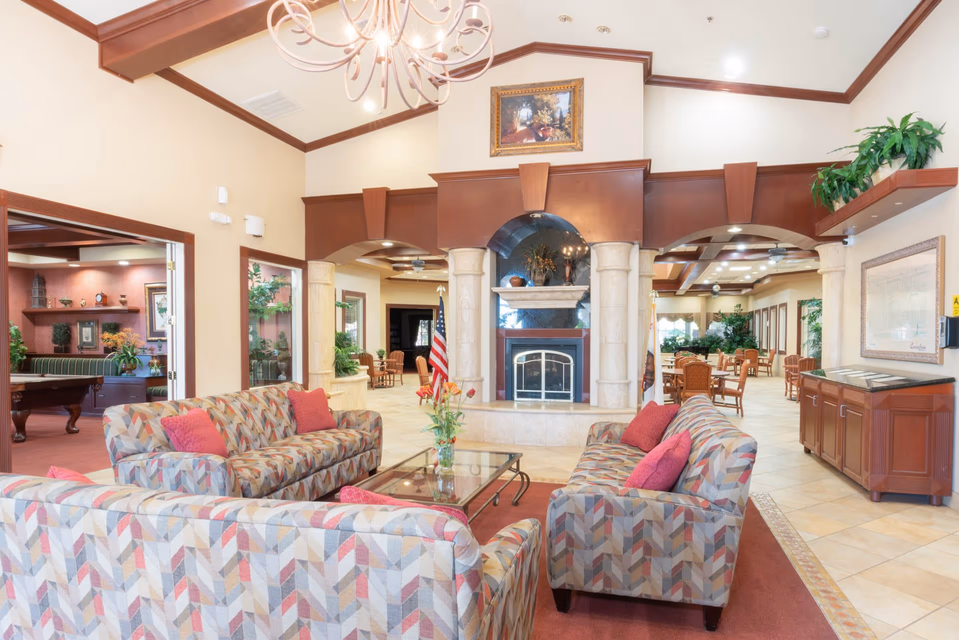 A spacious and well-lit living room area in a senior living facility with patterned sofas and red cushions arranged around a glass coffee table. The room features a fireplace with a decorative mantle, an American flag, and a chandelier hanging from the ceiling. In the background, there are dining tables and chairs, plants, and an adjacent room with a pool table and additional seating.