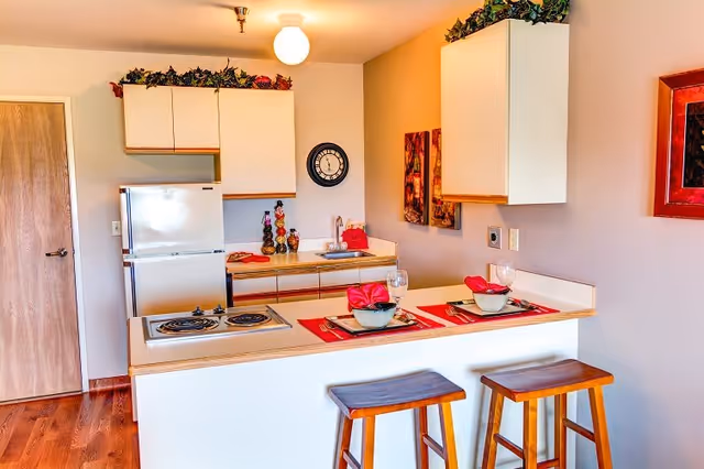 A small kitchen area with white cabinets and a white refrigerator. There is a countertop with two wooden stools and place settings for two, including bowls, glasses, and red napkins. The kitchen has a stove, a sink, and decorative items on the counter and above the cabinets. The walls are light-colored with a clock and framed artwork.