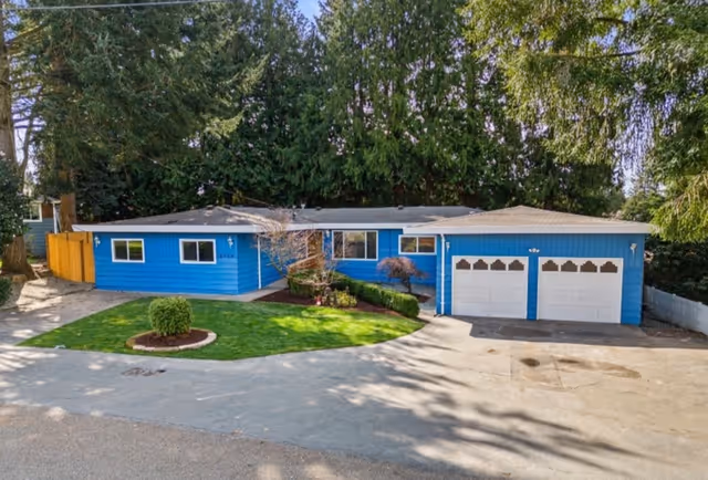 Single-story blue house with a two-car garage, surrounded by trees and a well-maintained lawn with a small circular garden bed in front.