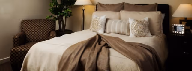 A cozy bedroom featuring a neatly made bed with beige and brown bedding, decorative pillows, and a brown throw blanket. To the left of the bed is a patterned armchair and a tall floor lamp with a beige shade. A small green plant is placed behind the armchair. On the right side of the bed is a dark wooden nightstand with a lamp and a framed photo.