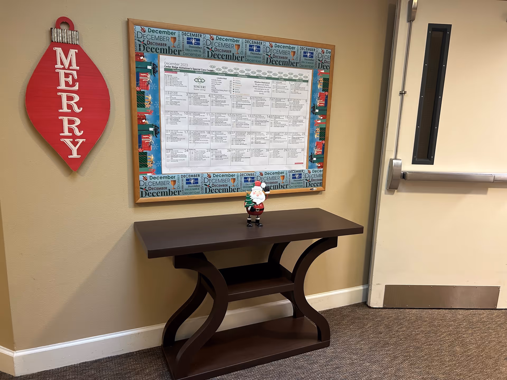 A hallway corner in Cedar Ridge Alzheimer's Special Care Center featuring a dark wooden console table with a small Santa Claus figurine on top. Above the table is a bulletin board with a December 2023 calendar and holiday-themed border. To the left of the bulletin board is a red wall decoration shaped like a Christmas ornament with the word 'MERRY' written vertically. To the right is a beige door with a narrow vertical window and metal kick plate.