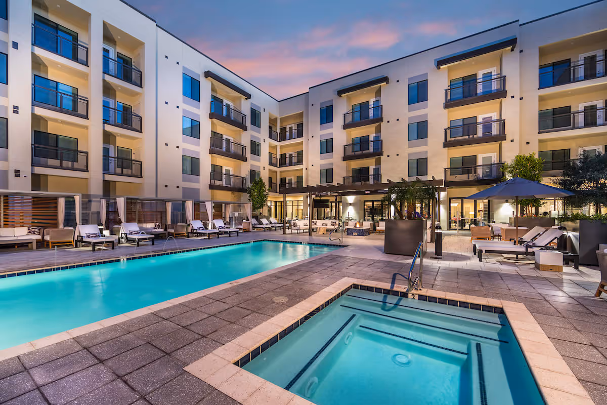 Courtyard pool and hot tub at a multi-story residential building with lounge chairs, umbrellas, and balconies at dusk.