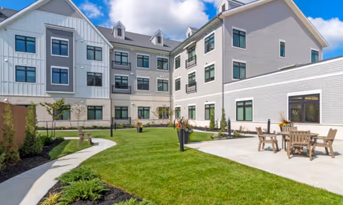 Well-maintained grassy courtyard with paved paths, patio seating and a modern three-story memory care building under a blue sky.