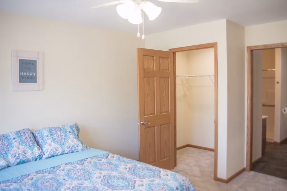 A bedroom with a bed covered in a blue and white patterned bedspread and matching pillows. There is a wooden door open to a walk-in closet with wire shelving. Another doorway leads to a bathroom. A ceiling fan with lights is visible above, and a framed sign on the wall reads 'Home is my Happy Place.'