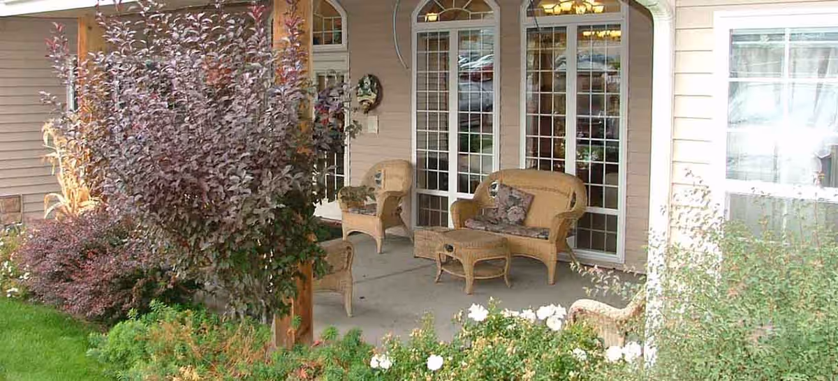 Outdoor patio area with wicker furniture including chairs, a loveseat, and a coffee table. The patio is surrounded by plants and bushes, and there are large windows and a door leading inside the building.