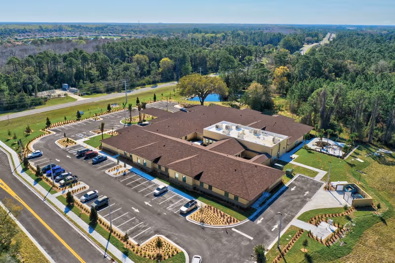 Aerial view of Gold Choice Palm Coast Assisted Living & Memory Care facility showing a single-story building with a brown roof surrounded by parking lots, landscaped areas, and greenery. The facility is located near a road with trees and open land in the background.
