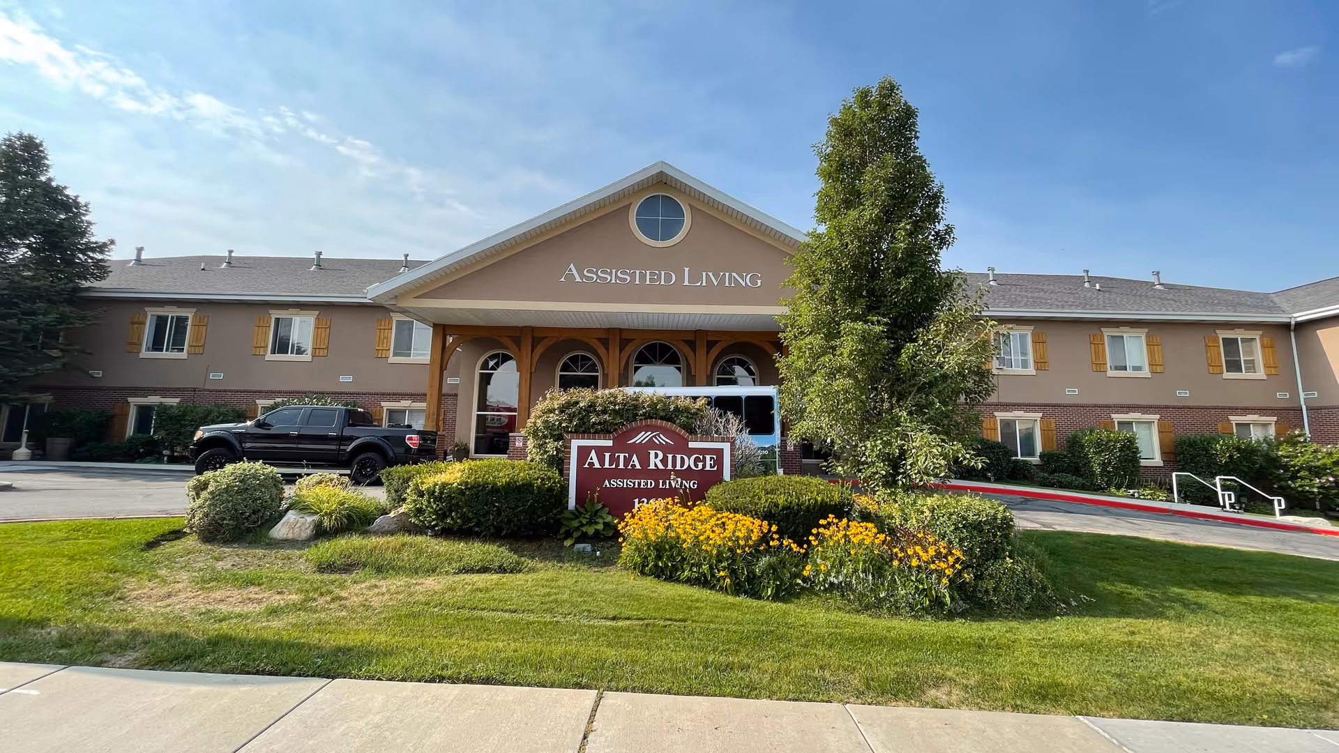 Front exterior view of Alta Ridge Assisted Living of Sandy, showing a two-story building with beige and brick facade, a covered entrance with wooden beams, a landscaped garden with yellow flowers and green bushes, and a sign reading 'Alta Ridge Assisted Living' in front.