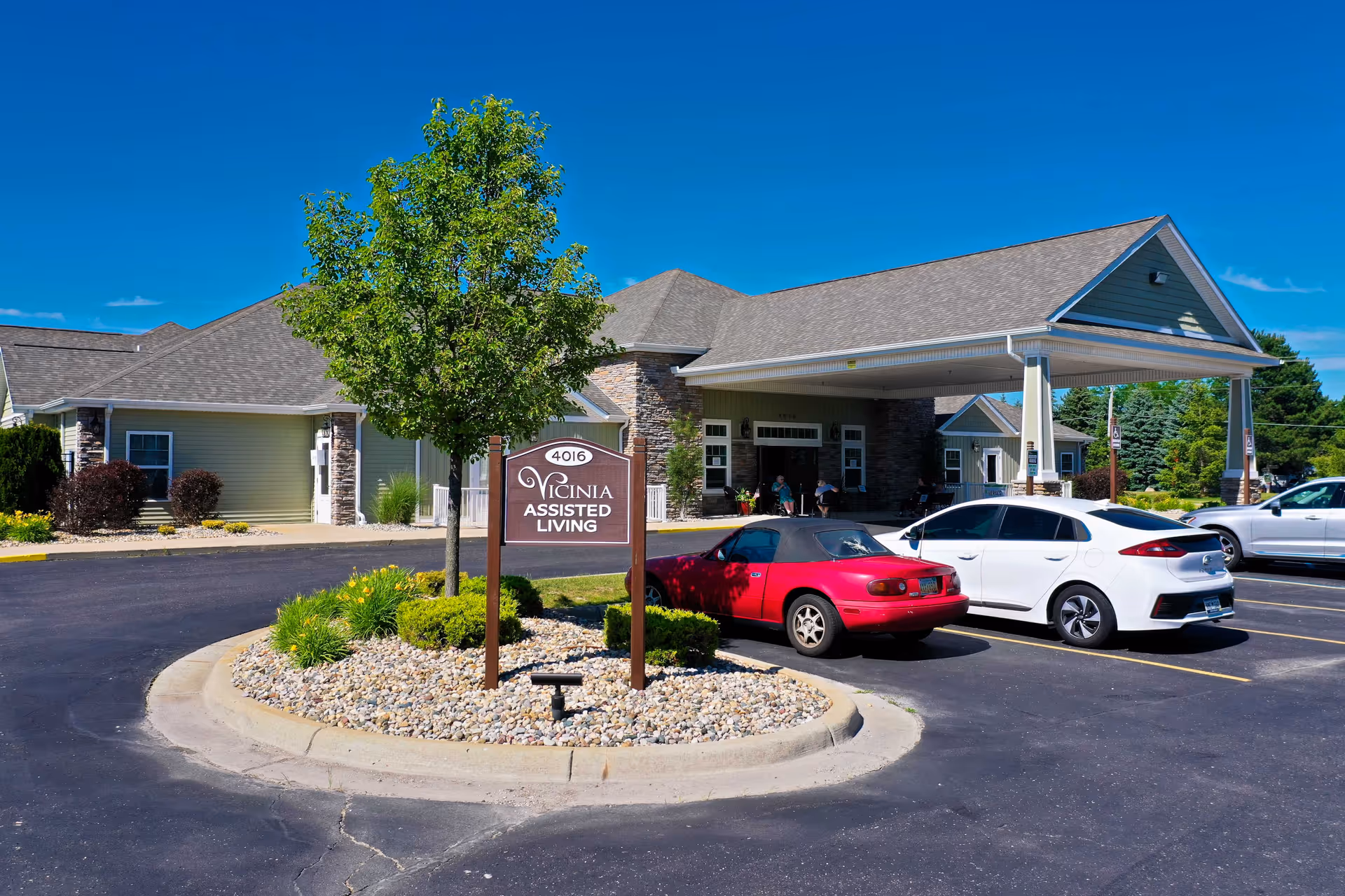 Exterior view of Vicinia Assisted Living facility showing the entrance with a covered drop-off area, a sign with the facility name, a small landscaped island with a tree and bushes, and several parked cars under a clear blue sky.