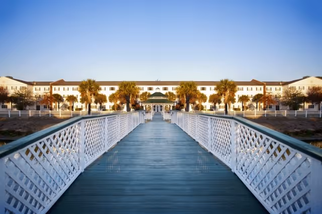 Long wooden walkway with white lattice railings leading to a large white building flanked by palm trees.