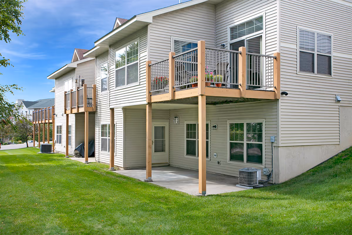 Exterior view of a residential building with beige siding, featuring multiple balconies with wooden railings and a well-maintained green lawn under a clear blue sky.
