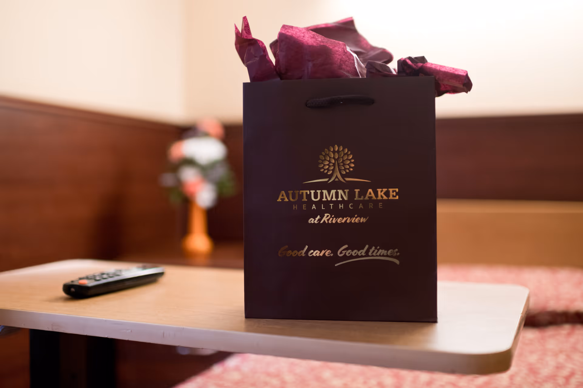 A dark gift bag with purple tissue paper sits on a light wooden table. The bag has the logo and text 'Autumn Lake Healthcare at Riverview' and the slogan 'Good care. Good times.' In the background, there is a blurred vase with flowers and a remote control on the table.