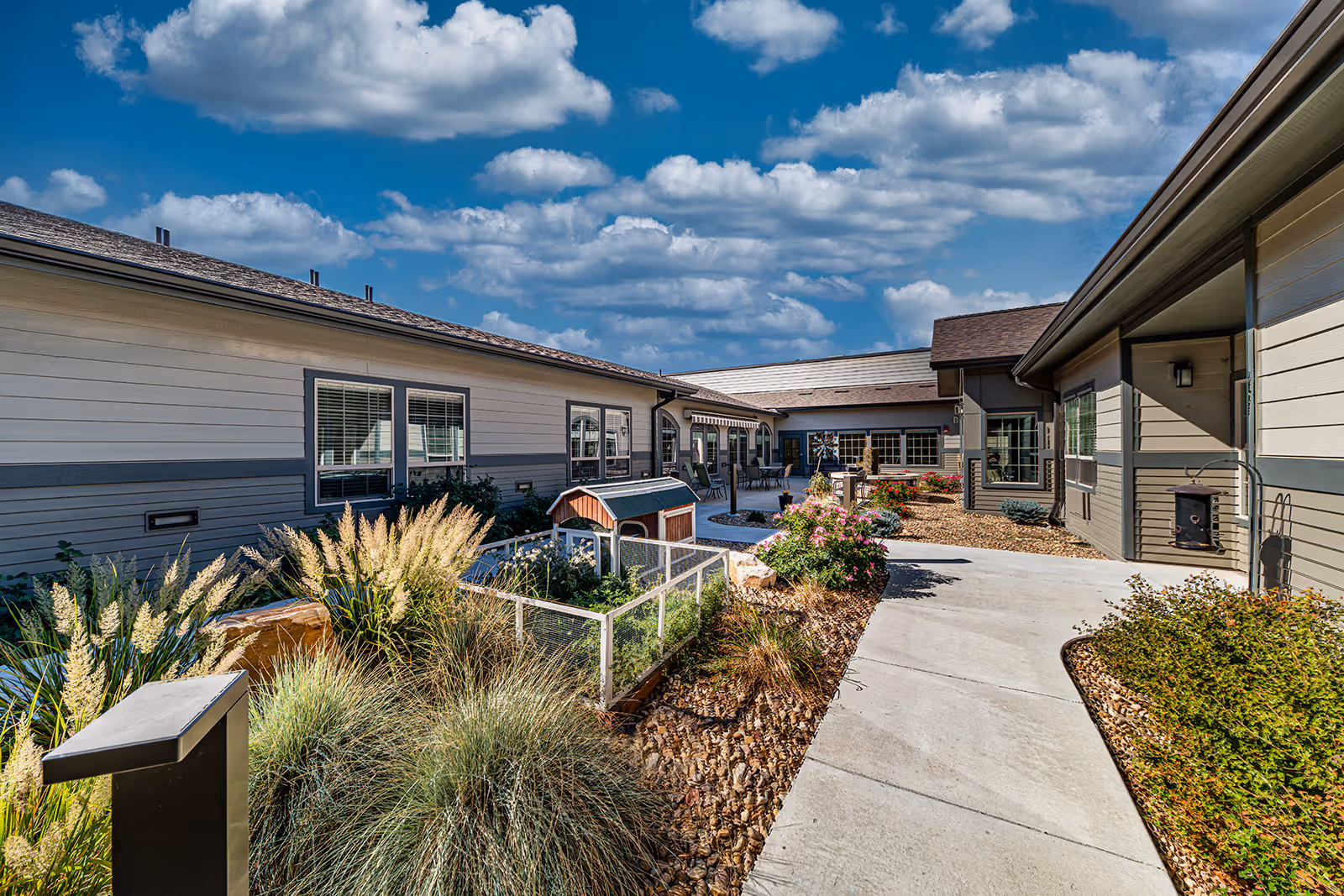 Outdoor courtyard area of a senior living facility with a paved walkway, landscaped garden beds containing ornamental grasses and flowering plants, and a small fenced garden with a wooden structure. The building surrounding the courtyard has beige and gray siding with multiple windows under a partly cloudy blue sky.