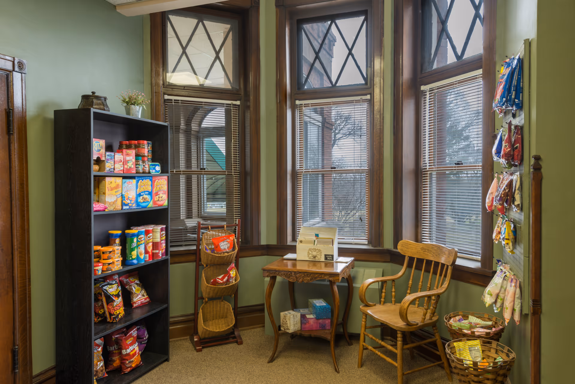 Small interior snack area with shelves of packaged snacks, a wooden chair and table by bay windows.