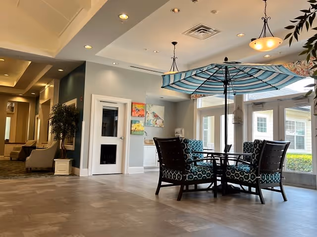 A bright and spacious interior common area with a round table and four cushioned chairs featuring a blue and white geometric pattern. Above the table is a large blue and white striped umbrella. The room has large windows letting in natural light, recessed ceiling lights, and a hallway with additional seating and plants visible in the background.