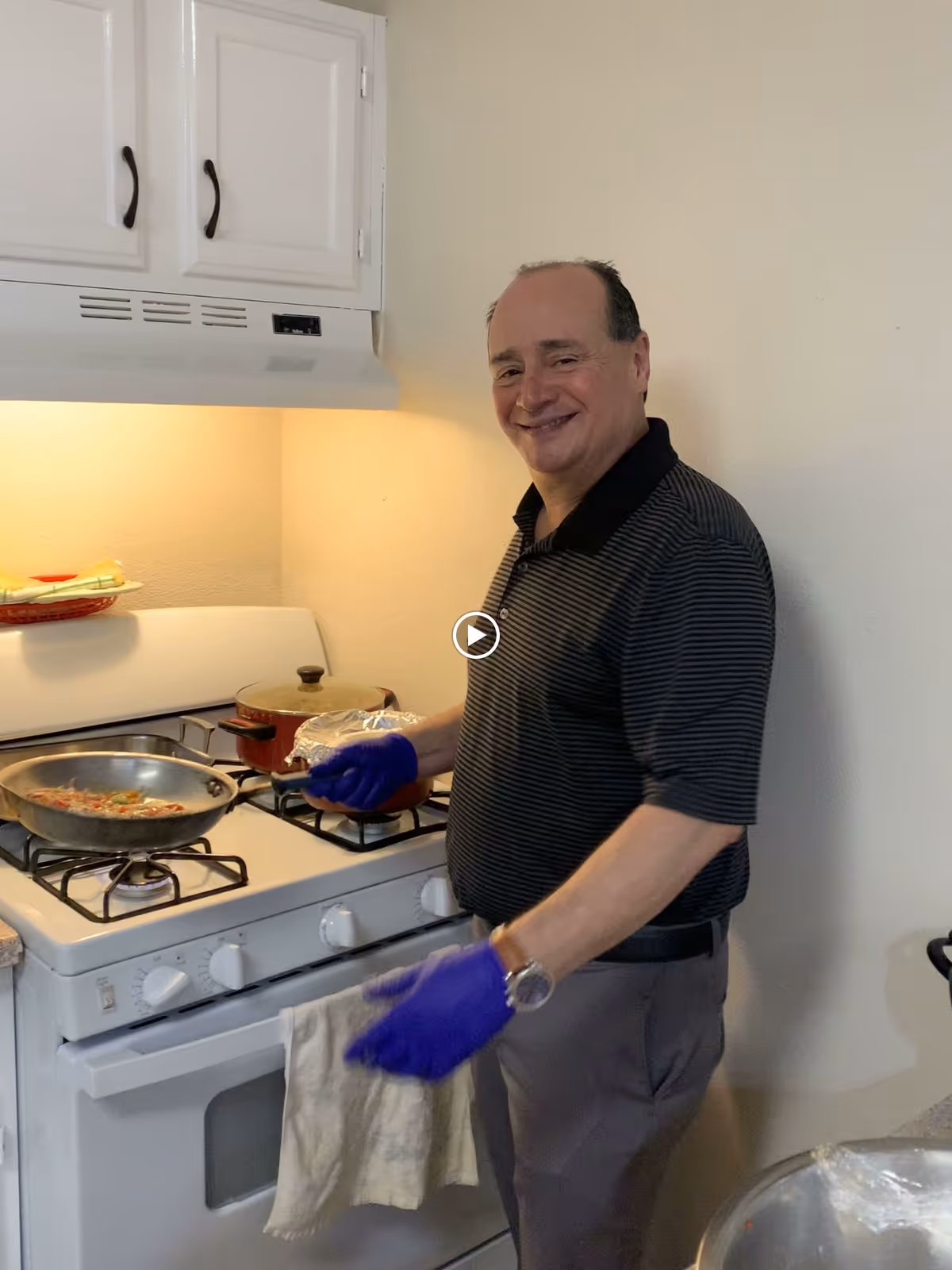 A man wearing a black and gray striped polo shirt and purple gloves is cooking on a white gas stove in a kitchen. There is a frying pan with food on one burner and a pot covered with foil on another burner. White cabinets and a range hood are visible above the stove.