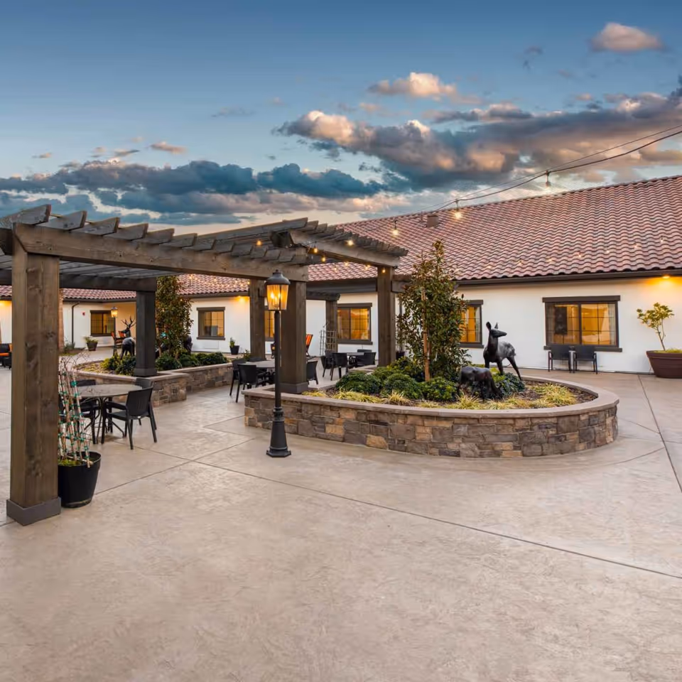 Outdoor courtyard area of a senior living facility with a wooden pergola, string lights, patio tables and chairs, landscaped garden beds with small trees and deer sculptures, and a building with a tiled roof and windows in the background under a partly cloudy sky.