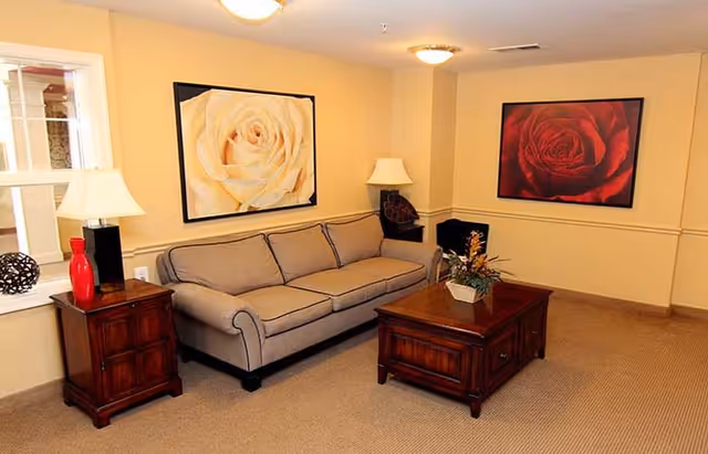 A cozy living room with a beige sofa, two framed rose paintings on the walls, a wooden coffee table with a floral arrangement, and two wooden side tables each with a lamp and decorative items.