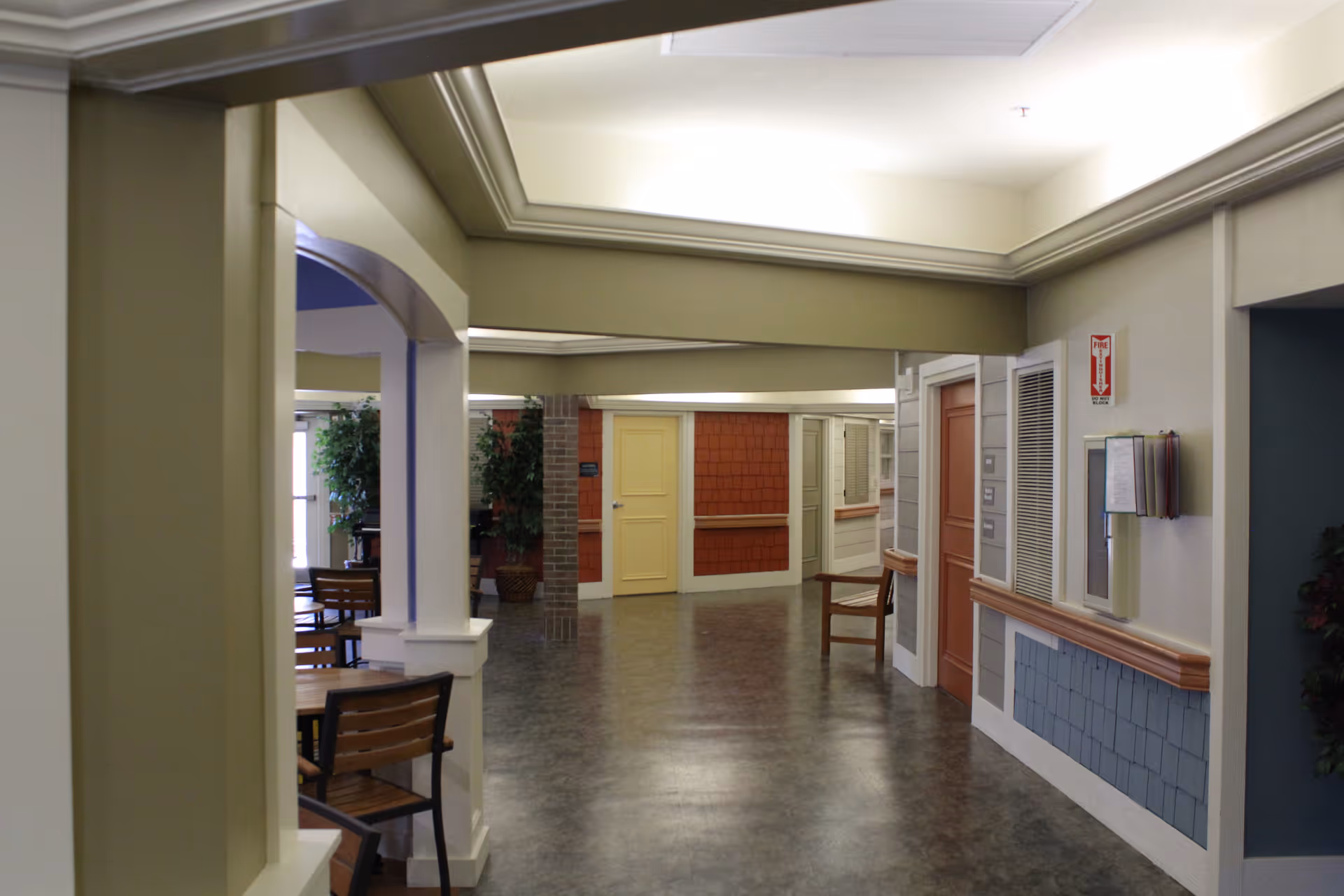 Interior hallway of a senior living facility with multiple doors, a chair, and some potted plants. The walls have decorative paneling and there is a fire extinguisher sign mounted on the wall.