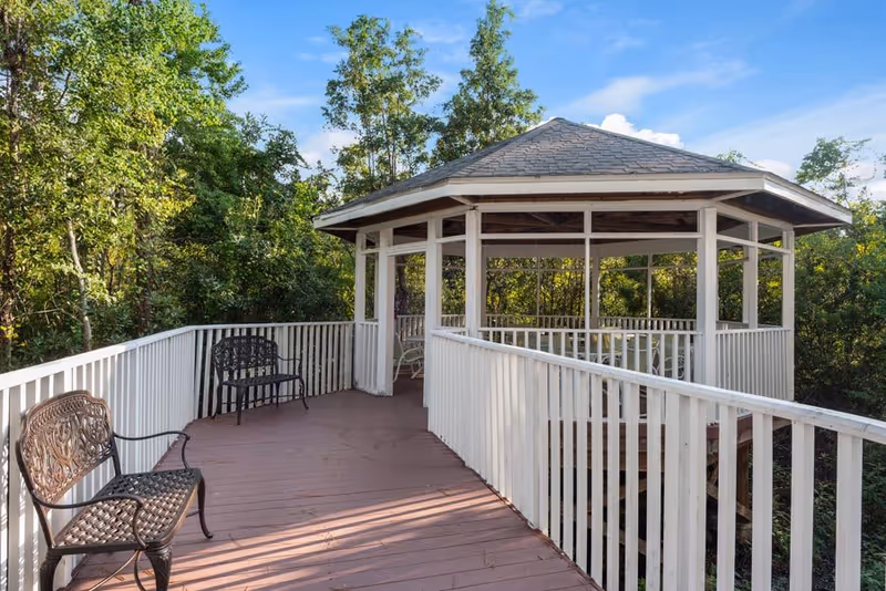 Outdoor wooden deck with white railings leading to a covered gazebo surrounded by trees under a blue sky. Two metal benches are placed along the deck.