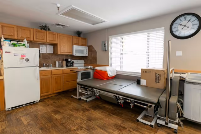 A room with a small kitchen area including a refrigerator, wooden cabinets, a microwave, a stove, and a coffee maker. Next to the kitchen is a medical bed or examination table with a red cushion and a cardboard box on it. A large window with blinds and a wall clock are visible on the right side.