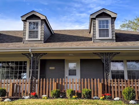 Front exterior of a single-story house with two dormer windows, a porch with decorative iron supports, a picket fence and a small flowerbed.