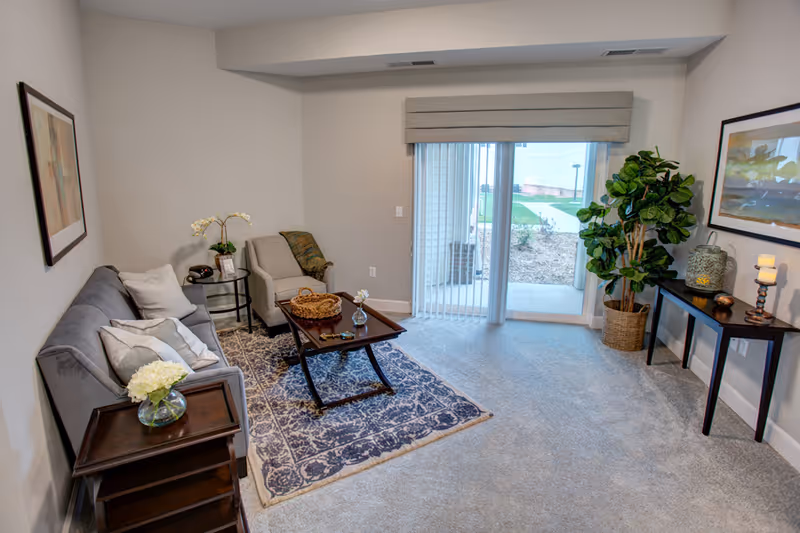 Neatly furnished living room with a sofa, armchair, coffee table on a patterned rug, and sliding glass doors to the outside.