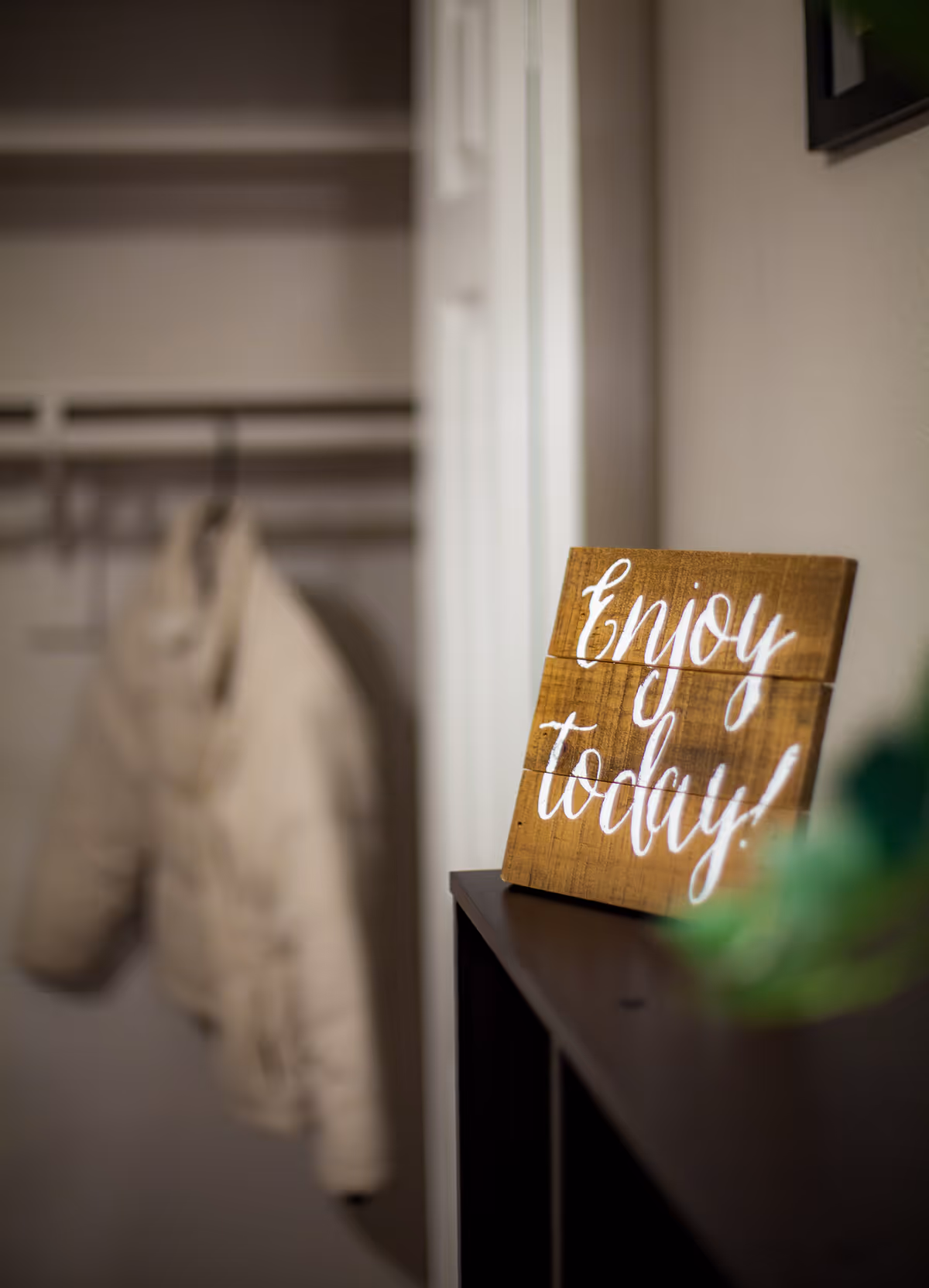A wooden sign reading "Enjoy today!" sits on a shelf with a blurred coat hanging in an open closet in the background.