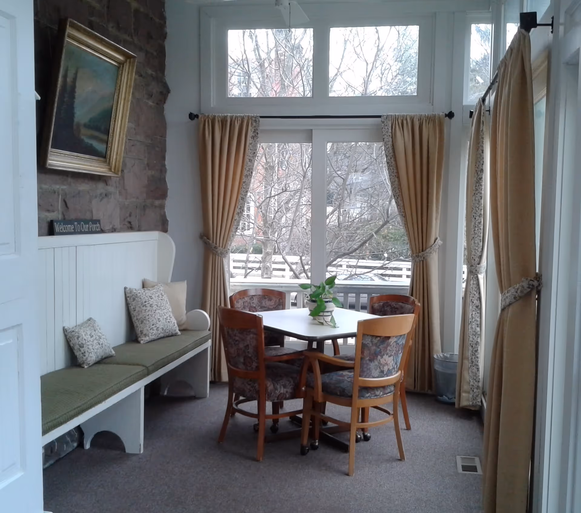 A cozy indoor seating area with a square table surrounded by four wooden chairs with floral upholstery. To the left is a built-in bench with green cushions and decorative pillows. Large windows with beige curtains let in natural light and show leafless trees outside. A framed painting hangs on a stone wall above the bench.