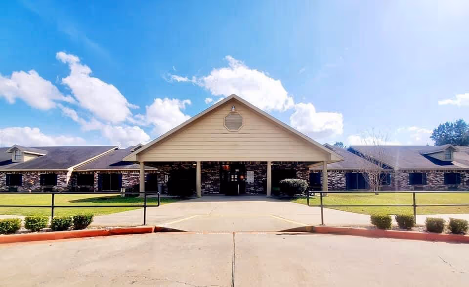 Front exterior view of Dogwood Trails Manor, a single-story building with a peaked roof entrance, brick walls, and a clear blue sky with some clouds in the background. There is a paved driveway and small bushes along the front.