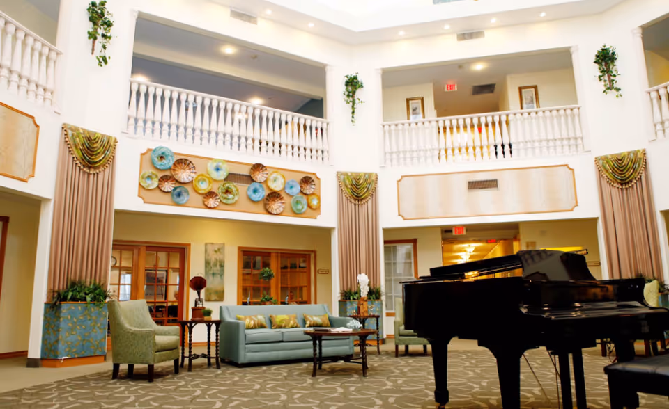 A spacious and elegant living room area in an assisted living facility featuring a grand piano, comfortable seating with a sofa and armchairs, decorative wall art, tall windows with drapes, and a high ceiling with a balcony railing on the second floor.
