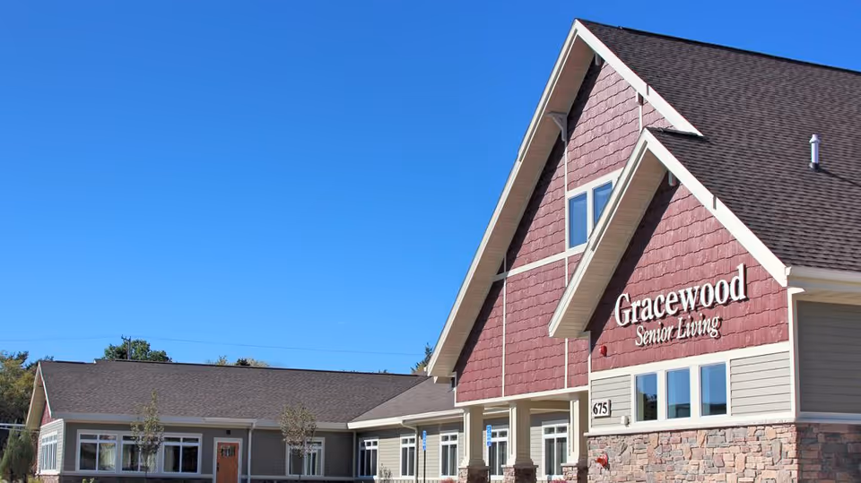 Exterior view of Gracewood Senior Living building with a clear blue sky. The building features a combination of red and beige siding with stone accents and multiple windows.