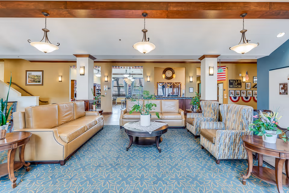 A bright and spacious senior living facility common area with beige leather sofas, patterned armchairs, wooden side tables with potted plants, and a round coffee table with a plant centerpiece. The room features a blue floral carpet, warm yellow walls, and a wooden ceiling beam with hanging light fixtures. In the background, there is a reception desk with a clock above it, and a 'Wall of Honor' display with American flags and framed photos.