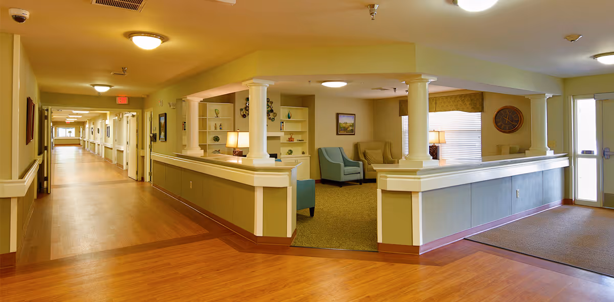 Interior view of a senior living facility hallway with wooden flooring and beige walls. On the right side, there is a common sitting area with armchairs, a lamp, wall art, and built-in shelves. The sitting area is partially enclosed by a half wall with white columns. The hallway extends into the distance with multiple doors and ceiling lights.