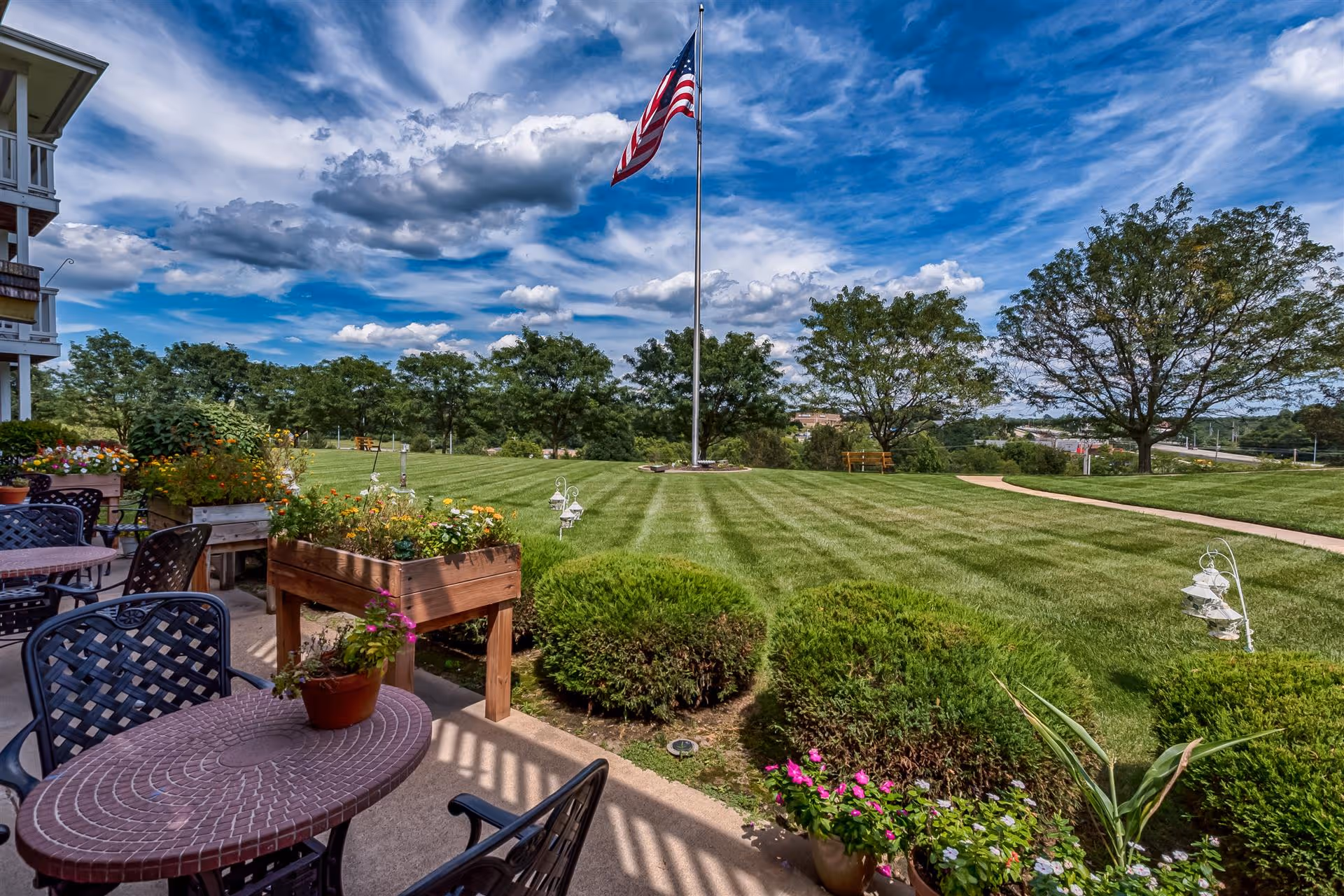 Outdoor patio area with round tables and chairs, potted plants, raised flower beds, and a well-maintained lawn with bushes and trees. An American flag flies on a tall flagpole in the center of the grassy area under a partly cloudy blue sky.