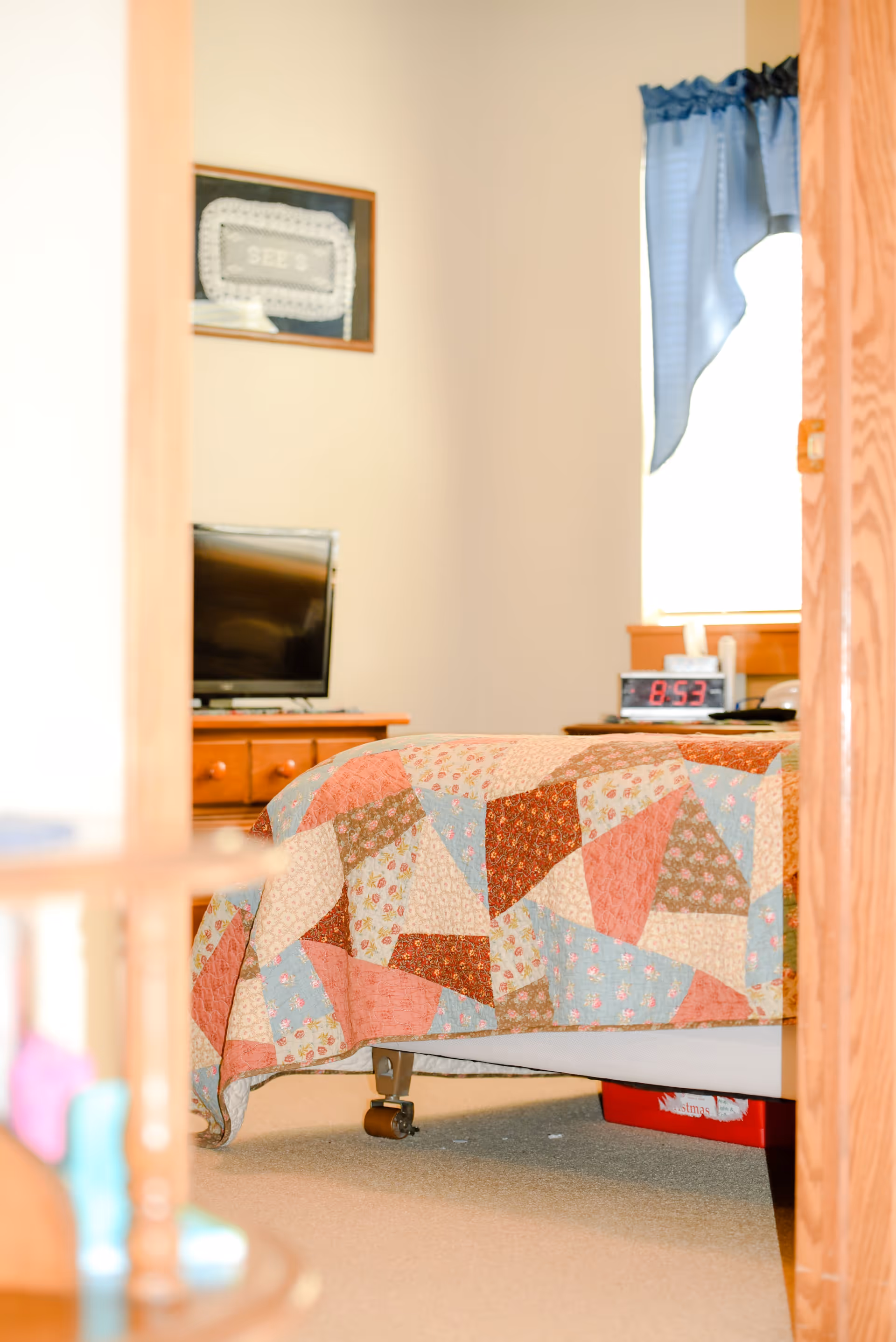 View through a doorway into a bedroom with a bed covered in a colorful patchwork quilt. A wooden dresser with a television on top is visible against the wall. A digital alarm clock showing 8:53 is on a nightstand near a window with blue curtains.