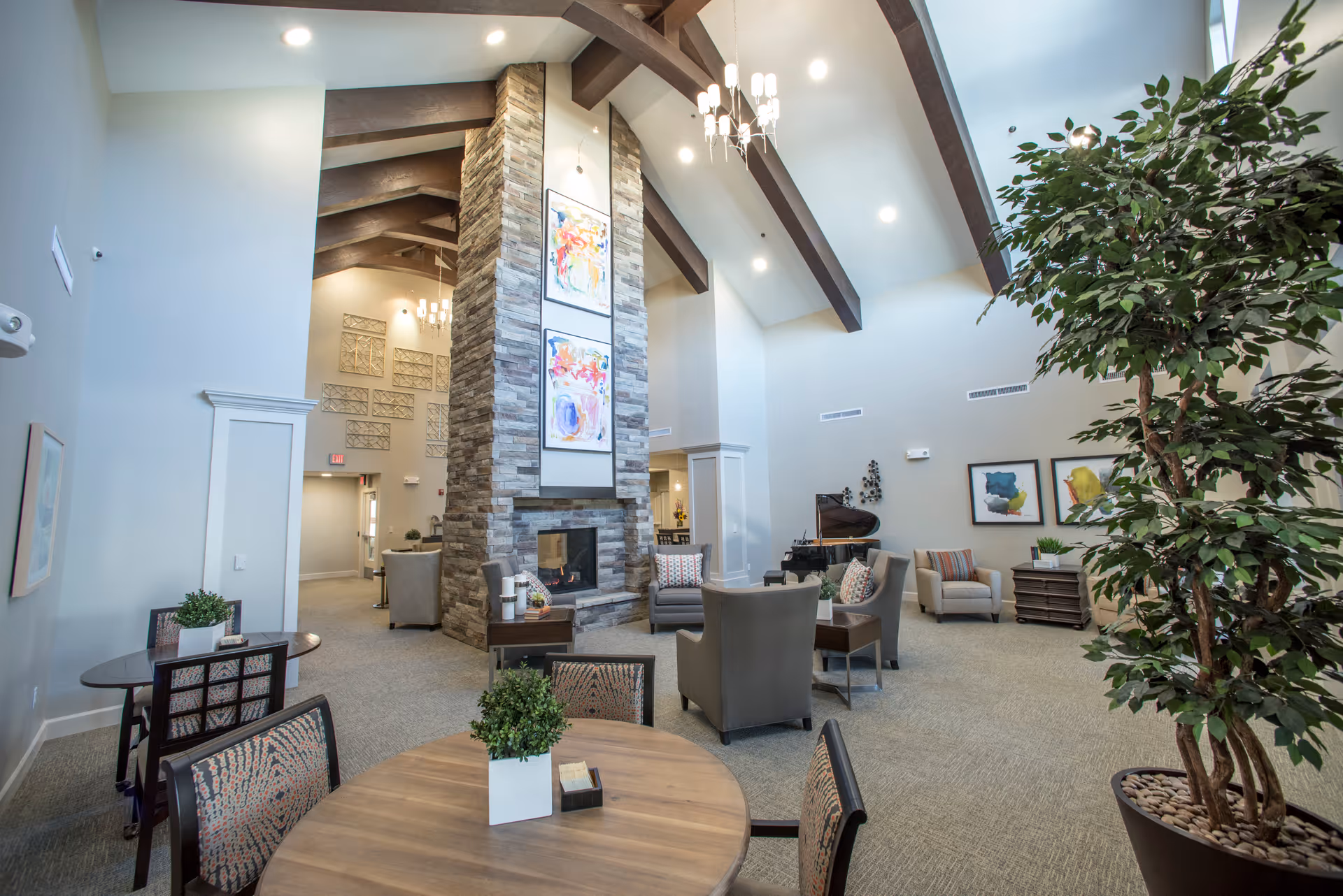 Spacious and well-lit common area with high ceilings and exposed wooden beams. The room features a large stone fireplace with colorful artwork above it, several comfortable armchairs arranged around small tables, a grand piano in the corner, and a large potted plant. There are also round tables with chairs and decorative plants, creating a welcoming and cozy atmosphere.