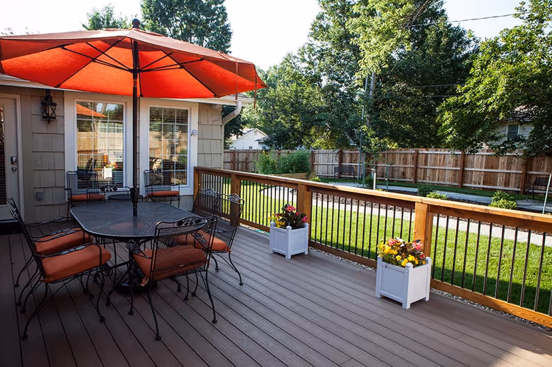 Outdoor wooden deck with a black metal table and six chairs with orange cushions under a large orange umbrella. Two white planters with colorful flowers are placed along the wooden railing. In the background, there is a fenced grassy yard with trees and a swing.