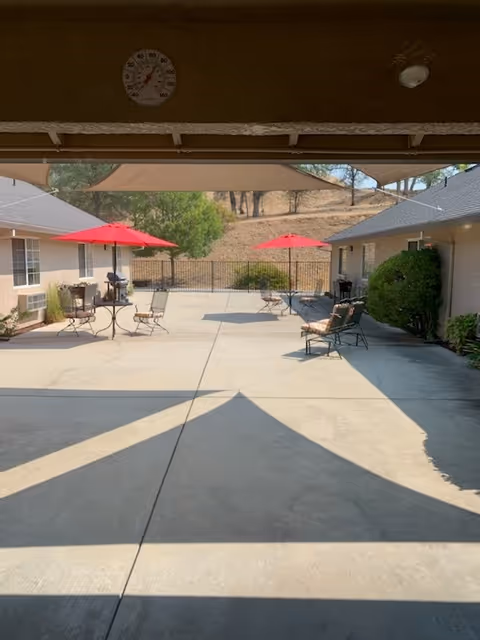 Outdoor courtyard area at Sierra Oaks Assisted Living & Memory Care with concrete flooring, several metal chairs and tables, and red umbrellas providing shade. The courtyard is surrounded by single-story buildings and has a backdrop of dry grassy hills and trees. A thermometer is mounted on the wall above the covered area.