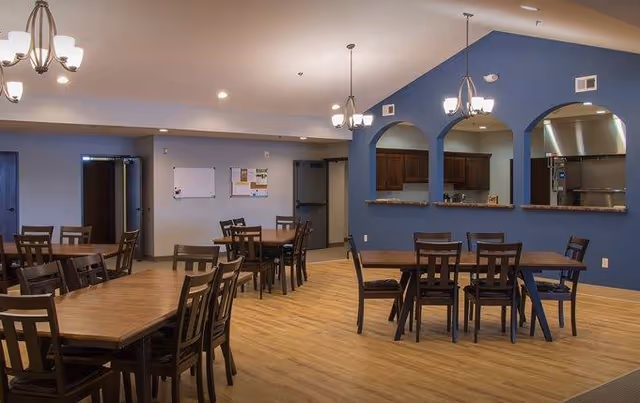 A spacious dining area with multiple wooden tables and chairs arranged neatly on a light wood floor. The room features a blue accent wall with three arched openings revealing a kitchen area behind it. Ceiling lights hang above the tables, and there are bulletin boards on the far wall.