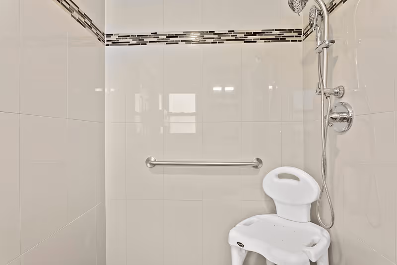 Tiled shower stall with a horizontal grab bar, handheld showerhead, and a white shower chair.
