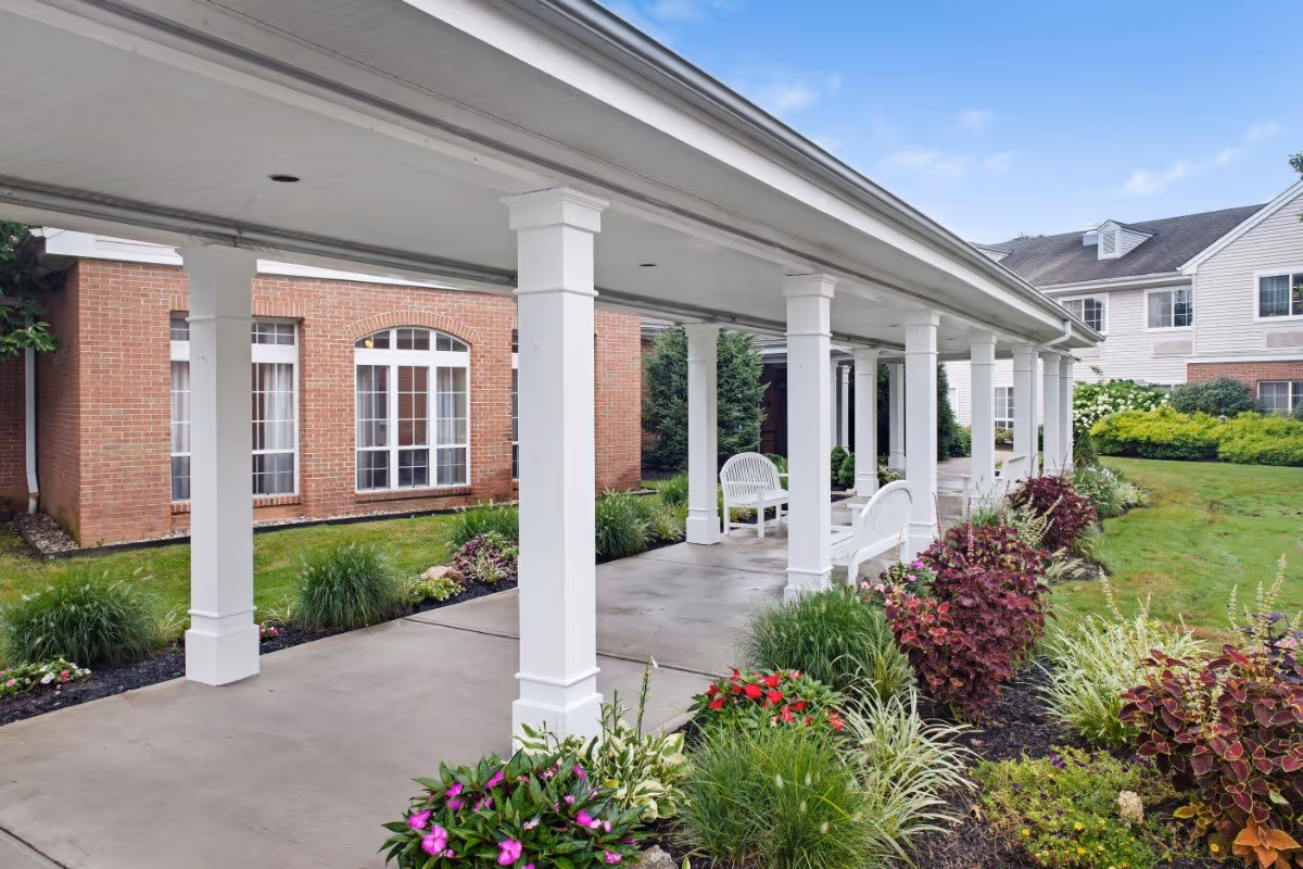 Covered outdoor walkway with white columns and benches, surrounded by landscaped garden beds with various plants and flowers, adjacent to a brick and siding building under a partly cloudy sky.