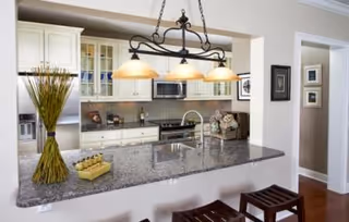 Modern kitchen area with a granite countertop island featuring a built-in sink and two wooden stools. The kitchen has white cabinets, a stainless steel refrigerator, a microwave, and decorative items including a vase with tall grass and a small tray with condiments.