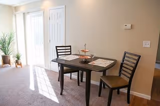 A small dining area with a dark wooden table set for two, featuring two chairs with cushioned seats, placemats, and a tiered serving tray. The room has beige walls, a thermostat on the wall, and a door leading outside with sunlight streaming in. There are two potted plants near the door.