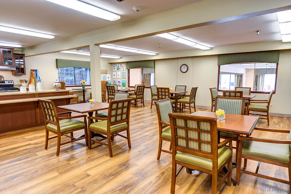 Bright communal dining room with wooden tables and green-upholstered chairs in a senior living facility.