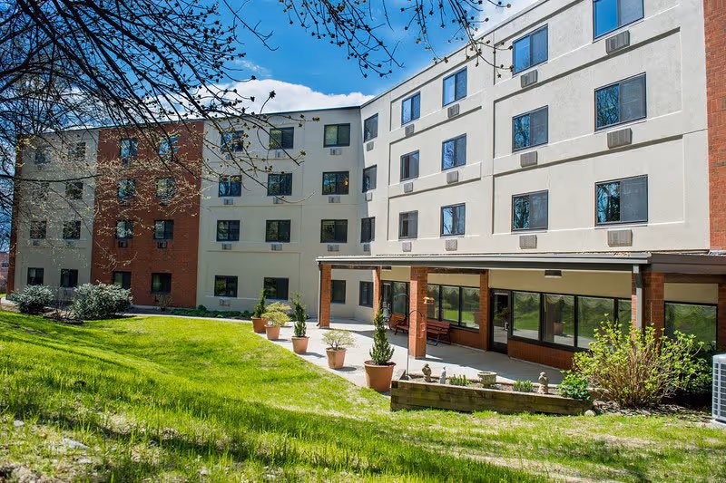 Exterior view of a multi-story senior living facility building with a combination of white and red brick walls, multiple windows, a covered patio area with benches, and a grassy lawn with potted plants and trees under a blue sky with some clouds.